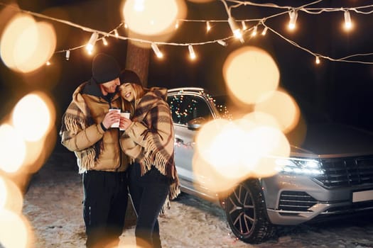 Standing near car. Couple is in the forest and celebrating New year.