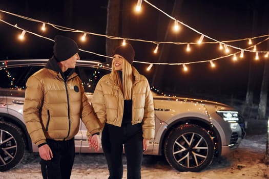 With modern car behind. Couple standing in the forest and celebrating New year.