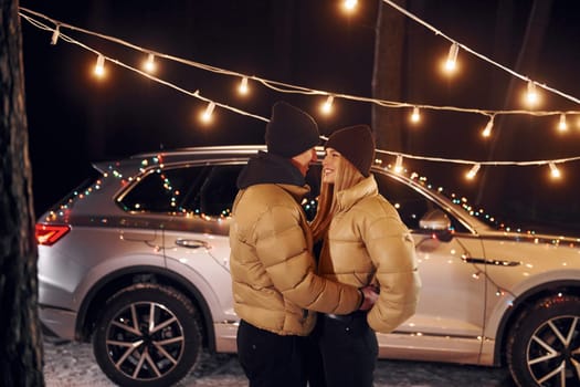 With modern car behind. Couple standing in the forest and celebrating New year.