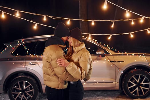 With modern car behind. Couple standing in the forest and celebrating New year.