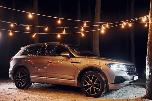 Modern silver colored automobile parked in forest with holiday decoration.