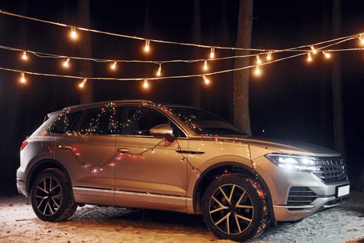 Modern silver colored automobile parked in forest with holiday decoration.