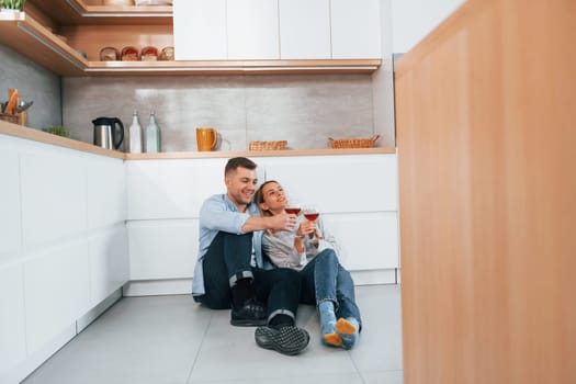 Drinking wine. Couple sitting on the floor of modern kitchen.