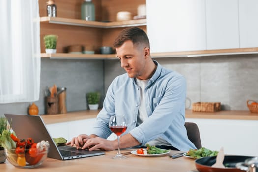 Using laptop. Man preparing food at home on the modern kitchen.