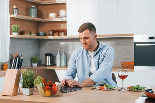 Using laptop. Man preparing food at home on the modern kitchen.