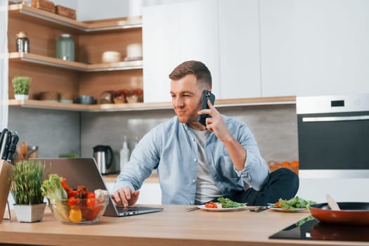 Talking by phone. Man preparing food at home on the modern kitchen.