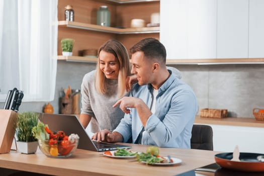 Looking at laptop. Couple preparing food at home on the modern kitchen.