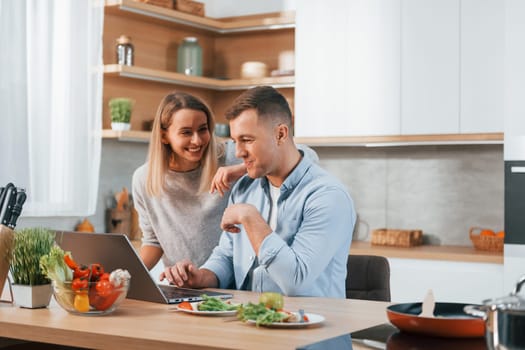 Looking at laptop. Couple preparing food at home on the modern kitchen.