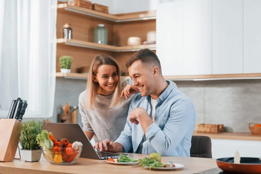 Looking at laptop. Couple preparing food at home on the modern kitchen.