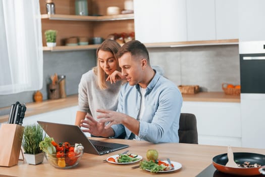 Looking at laptop. Couple preparing food at home on the modern kitchen.