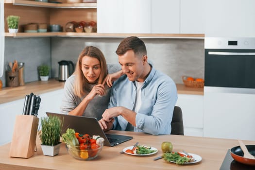 Laptop is on the table. Couple preparing food at home on the modern kitchen.