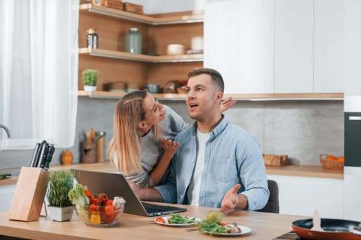 Laptop is on the table. Couple preparing food at home on the modern kitchen.