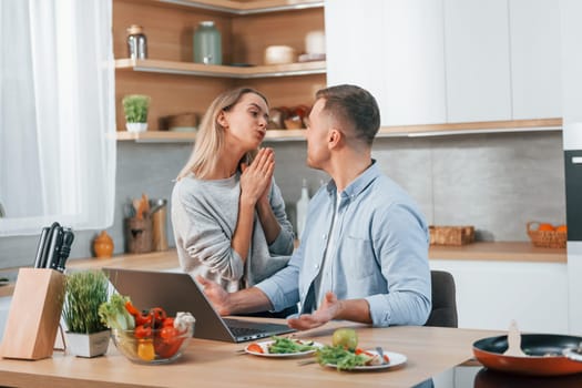 Laptop is on the table. Couple preparing food at home on the modern kitchen.