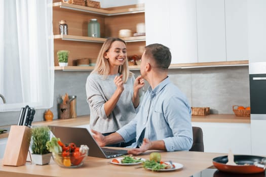 Laptop is on the table. Couple preparing food at home on the modern kitchen.