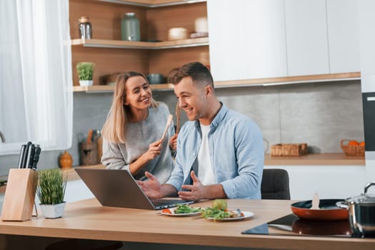 Modern laptop is on the table. Couple preparing food at home on the modern kitchen.
