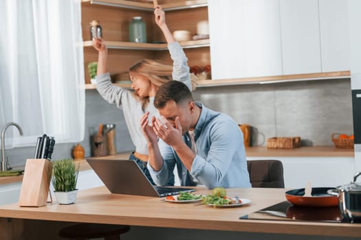 Modern laptop is on the table. Couple preparing food at home on the modern kitchen.