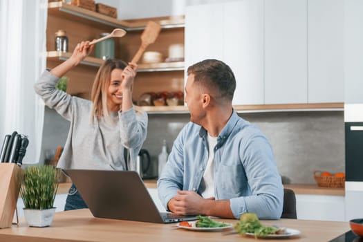 Modern laptop is on the table. Couple preparing food at home on the modern kitchen.