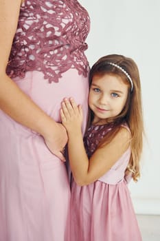 Conception of anticipation. Pregnant mother. Woman in dress standing with her daughter in the studio with white background.