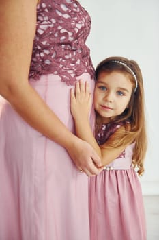 Woman is pregnant. Mother in dress standing with her daughter in the studio with white background.