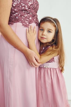 Woman is pregnant. Mother in dress standing with her daughter in the studio with white background.