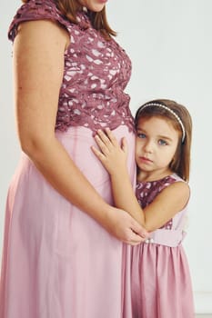Woman is pregnant. Mother in dress standing with her daughter in the studio with white background.