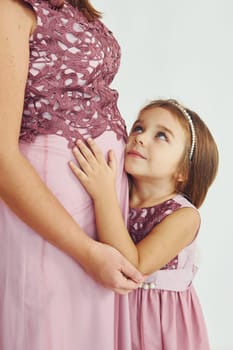 Woman is pregnant. Mother in dress standing with her daughter in the studio with white background.