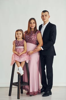 Girl is sitting on the chair. Happy couple with little daughter is standing in the studio.