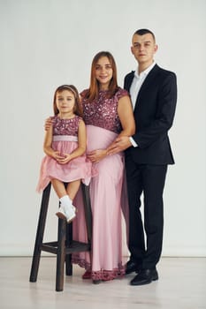 Girl is sitting on the chair. Happy couple with little daughter is standing in the studio.