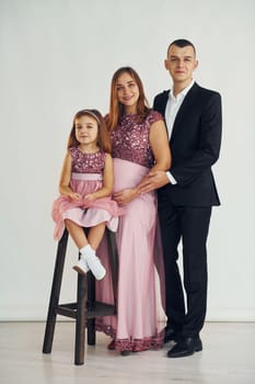 Girl is sitting on the chair. Happy couple with little daughter is standing in the studio.