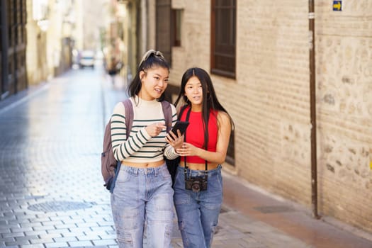 Young Asian female friends with backpacks and photo camera walking along street in Granada city while using map via mobile phone