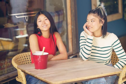 Smiling young Asian female friends in casual clothing sitting together at wooden table and discussing plans while spending time in cafe