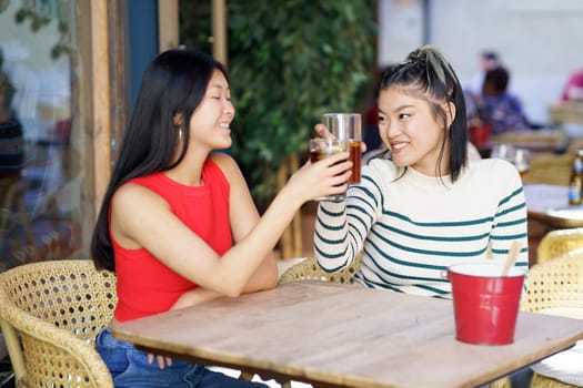 Delighted young Asian female friends, in casual outfits smiling and looking at each other while clinking glasses of drinks at wooden table in cafe during weekend together