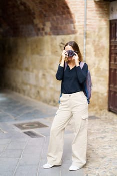 Full body of young female tourist in casual outfit with backpac,standing near aged brick building with arched passage and taking pictures on photo camera during sightseeing trip