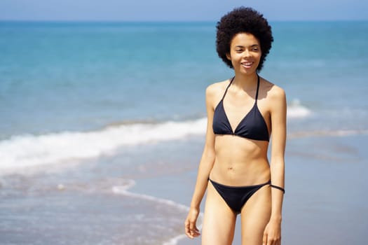 Cheerful African American female in swimwear looking into distance while strolling on coast along waving sea in tropical resort on summer day