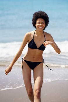 Delighted African American female in swimwear strolling on wet sandy beach near waving sea during summer vacation in tropical resort