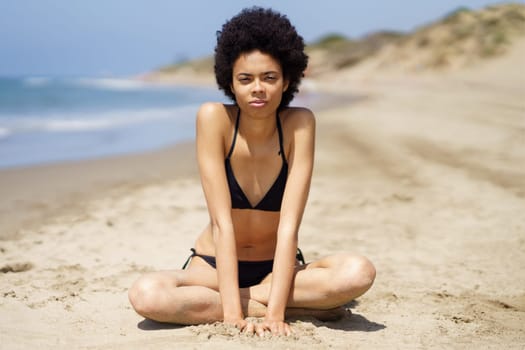 Full body of slim African American female traveler, in swimwear looking at camera with serious face, while sitting on sandy coast near sea during summer vacation