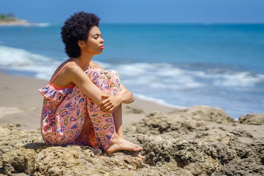 Barefoot black woman in ornamental maxi dress embracing knees and looking away while sitting on rough rocks against waving sea during summer vacation