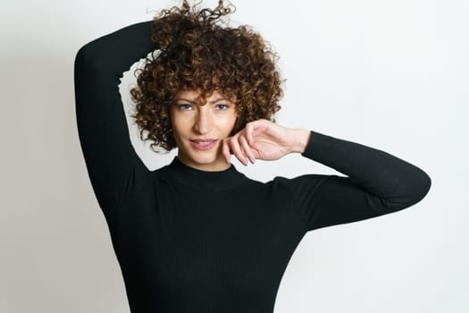 Positive girl model, with curly hair smiling and looking at camera while touching hands behind head against white background in unseen illuminating lights on shiny face