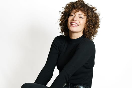 Positive young female with curly hair smiling with opened mouth and looking at camera while sitting with hands near leg against white background