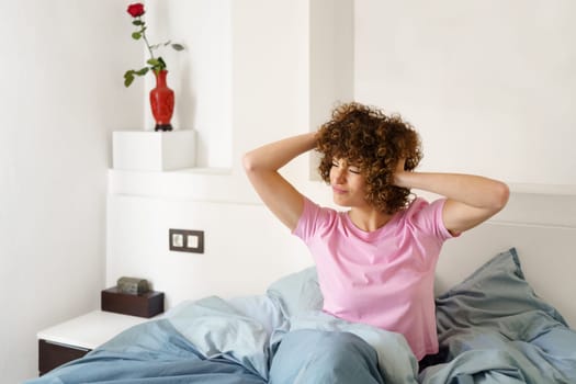 Curly haired female touching head and looking away while sitting on bed and getting up in morning