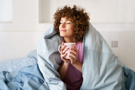 Relaxed curly woman with closed eyes wrapped in blanket while holding mug of hot beverage on bed in weekend