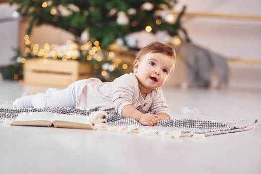 With book on the ground. Cute little baby is indoors in the domestic room.