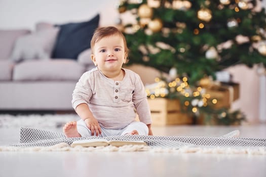 With book on the ground. Cute little baby is indoors in the domestic room.