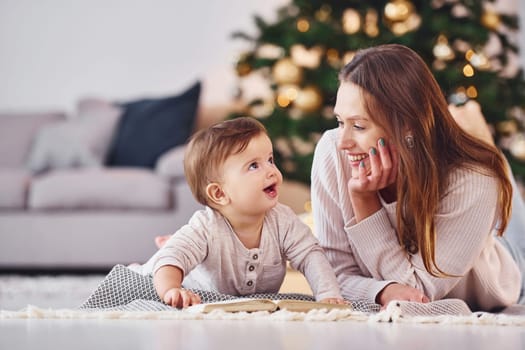 With book that is on the ground. Mother with her little daughter is indoors at home together.