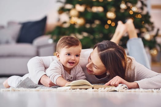 With book that is on the ground. Mother with her little daughter is indoors at home together.