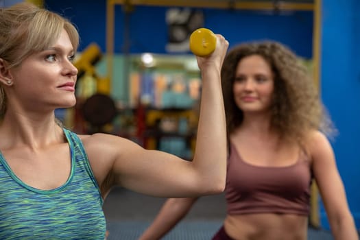 Two young and athletic girls are at the gym and one of them is lifting one dumbbell in her hand. Physical exercise for health and fitness.