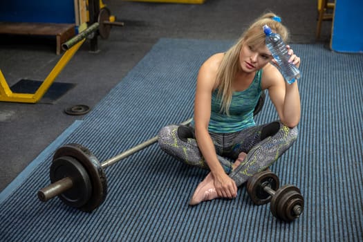 A tired girl sits on the carpet with her legs crossed. She puts a water bottle to her forehead. Behind the girl lies a barbell and in front of the girl lies an exercise weight.