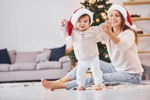 In Santa hats. Mother with her little daughter is indoors at home together.