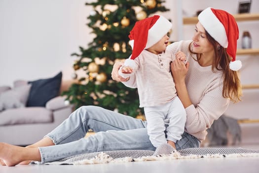 In Santa hats. Mother with her little daughter is indoors at home together.