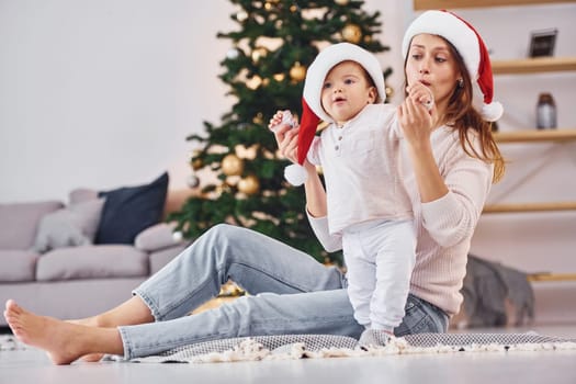 In Santa hats. Mother with her little daughter is indoors at home together.